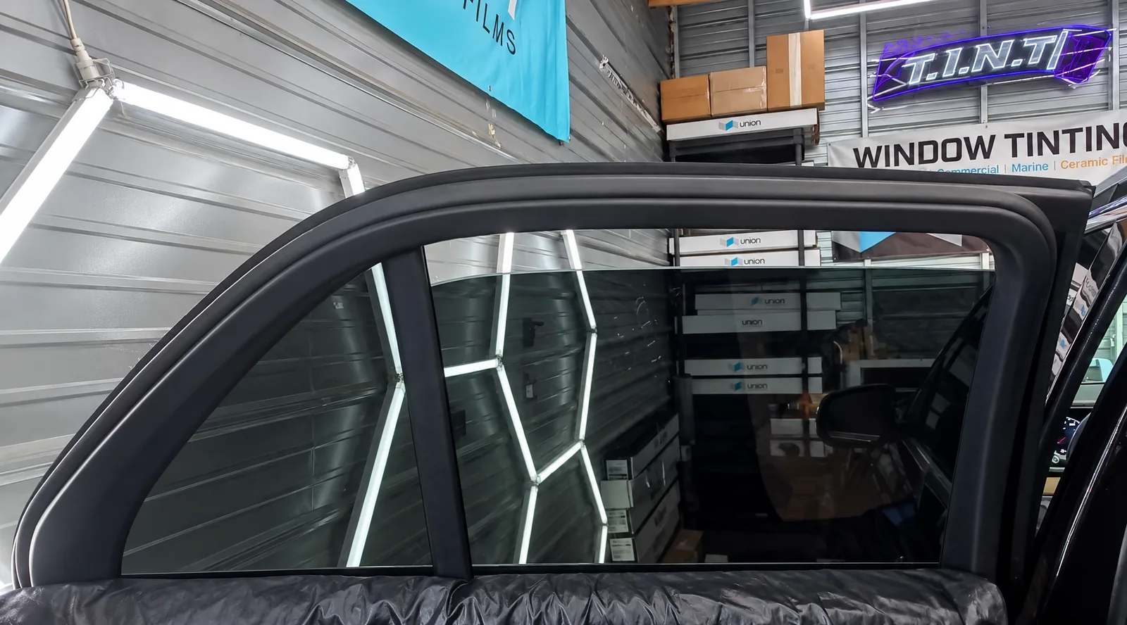 A rear door window freshly tinted in the bay, with the lit T.I.N.T sign on the wall behind